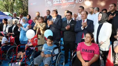 Arka planda "1001 Adet Medikal Malzeme Hoş Geldiniz" yazılı bir afişin önünde, tekerlekli sandalye kullanan çocuklar ve onları destekleyen çok sayıda kamu görevlisi ve vatandaş birlikte poz veriyor. Katılımcılar arasında takım elbiseli erkekler, kadınlar ve çocuklarla birlikte etkinliğe katılan gönüllüler bulunuyor. Topluluk alkış yapıyor.