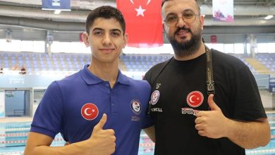 Görselde, bir yüzme havuzu önünde poz veren iki erkek yer alıyor. Soldaki genç sporcu, mavi renkte Türkiye ve federasyon amblemleri bulunan bir polo tişört giymiş, sağ baş parmağını kaldırarak gülümsüyor. Sağdaki kişi ise siyah tişört giymiş, üzerinde Türk bayrağı ve "Spor Toto" logosu yer alıyor. O da baş parmağını kaldırarak poz veriyor. Arka planda ise tribünler, Türk bayrağı ve yüzme havuzu görülüyor.