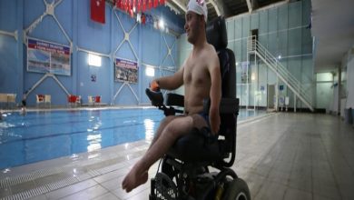 Görselde kapalı bir yüzme havuzunun kenarında tekerlekli sandalyede oturan genç bir sporcu bulunuyor. Başında bone var ve üst kısmı çıplak. Arka planda havuz, merdivenler, Türk bayrakları ve mavi duvarlı bir salon dikkat çekiyor. Sporcu gülümseyerek havuza doğru bakıyor.