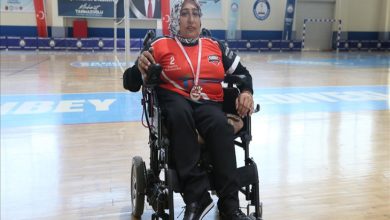 Görselde başörtülü Yeter Olcay, kırmızı ve siyah spor forması giymiş halde tekerlekli sandalyesinde oturuyor. Boynunda madalya asılı. Spor salonunun ortasında duruyor ve arka planda pankartlar ile Türk bayrağı görünüyor.