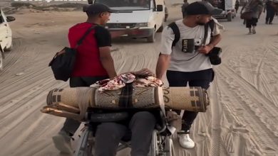 Görselde, kumlu bir alanda kalabalık bir grup insan görülüyor. Ön planda bir kişi tekerlekli sandalyede, üzerine eşyalar (halı, torba vb.) yüklenmiş şekilde taşınıyor. Yanında iki kişi sandalyeyi iterek yardım ediyor. Arka planda araçlar ve yürüyen insanlar bulunuyor. Görselin sol üst köşesinde “Yeni Şafak” logosu yer alıyor.