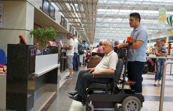 Görselde bir havalimanı terminalinde yolcular işlem sırası bekliyor. Ön planda yaşlı bir yolcu tekerlekli sandalye ile görevli bir personel tarafından bankoya götürülüyor. Arkada ise check-in işlemleri için sırada bekleyen başka yolcular görünüyor. Ortam kalabalık ve hareketli.