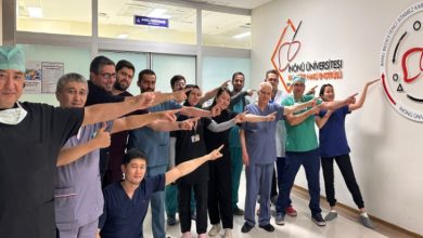 Görselde, ameliyathane kapısının önünde bir grup sağlık çalışanı (doktorlar, hemşireler ve diğer personel) poz veriyor. Hepsi aynı yöne, duvarda yer alan “İnönü Üniversitesi Karaciğer Nakli Enstitüsü” logosuna parmaklarıyla işaret ediyor. Gruptaki herkes mutlu ve gururlu görünüyor.