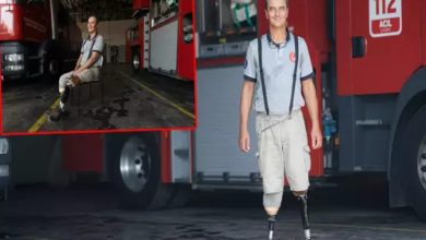 Görselde, bir itfaiyeci üniforması giymiş, protez bacakları olan bir kişi itfaiye aracının önünde gülümseyerek ayakta poz veriyor. Arka planda kırmızı itfaiye aracı dikkat çekiyor. Sol üst köşede aynı kişinin itfaiye garajında sandalyede otururken çekilmiş başka bir fotoğrafı da küçük bir çerçeve içinde yer alıyor.