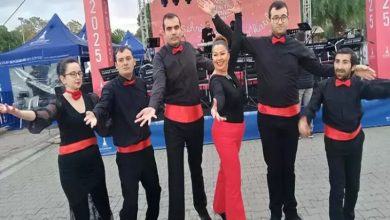 Görselde altı kişilik bir dans ekibi sahne önünde poz veriyor. Hepsi siyah kıyafetler giymiş, kırmızı kuşak ve papyon takmış. Ortam açık hava etkinliğini andırıyor; sahnede müzik ekipmanları ve arka planda etkinlik afişi var.