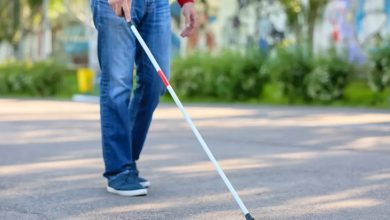 Görselde, açık bir alanda yürüyen görme engelli bir bireyin alt gövdesi görülüyor. Kişi, bir elinde beyaz baston tutarak yolda ilerliyor. Üzerinde kot pantolon ve kırmızı bir üst bulunmakta. Bastonun ucu yere temas etmiş durumda, bu da bireyin yönünü ve yolunu bu baston aracılığıyla belirlediğini gösteriyor. Arka planda yeşillikler ve hafif flu bir çevre manzarası var.