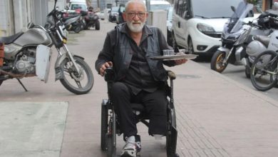 Görselde, akülü tekerlekli sandalye kullanan yaşlı bir adam sokakta ilerlerken görülüyor. Elinde çay bardakları bulunan bir tepsi taşıyor. Arka planda motosikletler, arabalar ve kaldırım üzerindeki yol detayları yer almakta. Adamın yüzünde hafif bir tebessüm var.