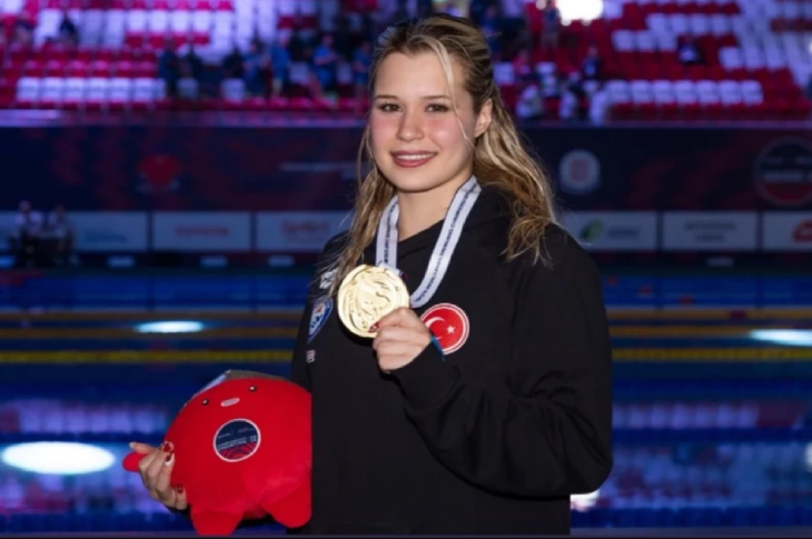 Milli para yüzücü Defne Kurt, şampiyona maskotunu sağ elinde tutarken boynunda duran altın madalyayı sol eliyle tutup gülerek poz veriyor.