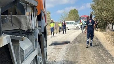 Görselde kırsal bir yolda trafik kazası sonrası olay yeri görüntülenmiş. Ön planda bir asfalt kamyonu, arka planda ise polis, sağlık ekipleri ve bir beyaz araç görülüyor. Yerde bir kişinin üzeri bulanıklaştırılmış. Olay yerinde güvenlik güçleri ve sağlık görevlileri inceleme yapıyor.