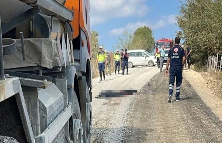 Görselde kırsal bir yolda trafik kazası sonrası olay yeri görüntülenmiş. Ön planda bir asfalt kamyonu, arka planda ise polis, sağlık ekipleri ve bir beyaz araç görülüyor. Yerde bir kişinin üzeri bulanıklaştırılmış. Olay yerinde güvenlik güçleri ve sağlık görevlileri inceleme yapıyor.
