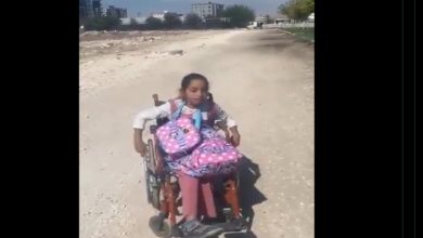 Görselde, okul çantasıyla tekerlekli sandalyede ilerlemeye çalışan küçük bir kız çocuğu, bozuk ve taşlık bir yolda görülüyor. Arka planda yerleşim yerleri bulunmakta. Kare, erişilebilir olmayan yolların engelli çocuklar için nasıl bir zorluk yarattığını çarpıcı şekilde yansıtıyor.