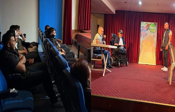 Görselde bir tiyatro salonu sahnesi ve izleyiciler görülüyor. Sağ tarafta sahnede iki masa, üzerinde oturan kişilerden biri tekerlekli sandalyede, diğeri ise masada oturuyor. Yanlarında ayakta duran bir oyuncu daha var. Arka fonda kırmızı perde ve üzerinde “SİBAS” yazılı renkli bir afiş bulunuyor. Sol tarafta ise salonda oturan izleyiciler oyunu dikkatle takip ediyor.