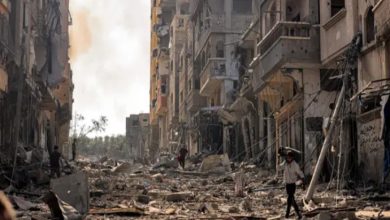 Görselde, savaşın harabeye çevirdiği bir şehir manzarası yer alıyor. Her iki yanda yüksek binalar ağır hasar almış, cepheleri yıkılmış, camlar kırılmış ve enkaz yığınları sokağa saçılmış durumda. Sokakta, yıkıntıların arasında yürümeye çalışan birkaç sivil insan görülüyor; biri başını korur şekilde elini yukarı kaldırmış. Elektrik direkleri devrilmiş, kablolar yerlere sarkmış. Gökyüzü dumanlı, ortam gri ve kasvetli. Bu kare, Gazze’de yaşanan yıkımın boyutunu ve sivil halkın çaresizliğini çarpıcı bir şekilde gözler önüne seriyor.