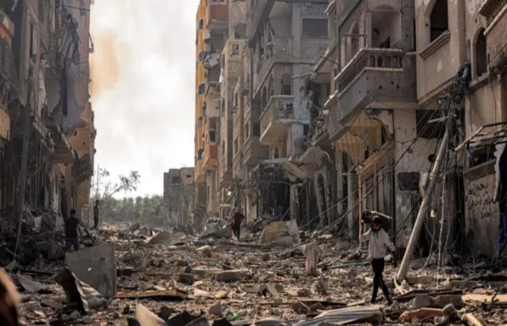 Görselde, savaşın harabeye çevirdiği bir şehir manzarası yer alıyor. Her iki yanda yüksek binalar ağır hasar almış, cepheleri yıkılmış, camlar kırılmış ve enkaz yığınları sokağa saçılmış durumda. Sokakta, yıkıntıların arasında yürümeye çalışan birkaç sivil insan görülüyor; biri başını korur şekilde elini yukarı kaldırmış. Elektrik direkleri devrilmiş, kablolar yerlere sarkmış. Gökyüzü dumanlı, ortam gri ve kasvetli. Bu kare, Gazze’de yaşanan yıkımın boyutunu ve sivil halkın çaresizliğini çarpıcı bir şekilde gözler önüne seriyor.