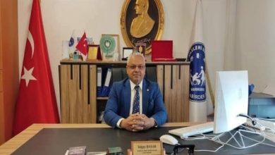 Görselde bir makam odasında oturan takım elbiseli bir erkek yer alıyor. Masanın üzerinde bilgisayar, evraklar ve bir isimlik bulunuyor. Arka planda Atatürk portresi, Türk bayrağı ve engellilerle ilgili bir derneğin bayrağı dikkat çekiyor. Bu ortam, resmi bir toplantı ya da kurum odasını yansıtıyor.