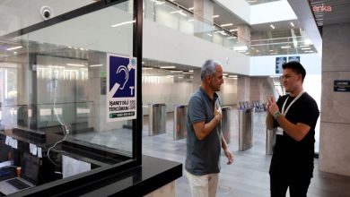 Görselde, modern bir kamu binasının girişinde iki kişi işaret diliyle iletişim kuruyor. Biri gri tişört, diğeri siyah tişört giymiş. Yan tarafta camlı bir danışma bölmesi ve üzerinde işitme engelliler için “İşaret Dili Tercümanı Hizmeti” yazılı bir afiş yer almakta. Arkada turnikeler ve geniş, ferah bir bina içi görülüyor.