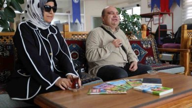 Geleneksel motiflerle süslenmiş bir oturma alanında görme engelli yazar Selman Devecioğlu ile eşi yan yana oturuyor. İkisi de gülümseyerek sohbet ederken, önlerindeki masada birkaç kitap ve çay bardağı bulunmaktadır. Arka planda ise ofis ya da sosyal tesis havası veren bir iç mekân görünmekte, duvarlarda çeşitli bayraklar ve afişler yer almaktadır.