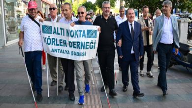 Görselde bir grup kişi kaldırımda yürüyüş yapıyor. Ön sırada “Altı Nokta Körler Derneği Antalya Şubesi” yazılı bir pankart taşıyan kişiler var. Bazı katılımcılar beyaz baston kullanıyor. Arka planda diğer yürüyüşçüler ve şehir ortamı görülüyor. Katılımcılar gülümseyerek birlikte ilerliyor.