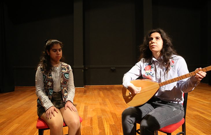 Görselde sahne üzerinde iki genç kadın oturuyor. Sağda oturan kadın bağlama çalıyor, soldaki ise gözleri kapalı şekilde dinliyor. Her ikisi de sahne ışığı altında kırmızı sandalyelerde oturmakta. Arka plan siyah, zemin ise ahşap kaplamalı. Ortam bir müzik performansı ya da prova alanını andırıyor.