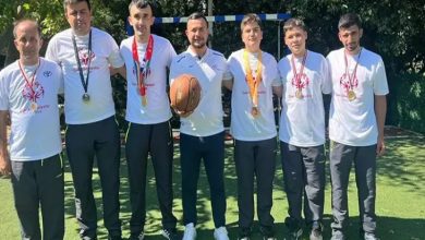 Görselde, açık havada bir araya gelmiş bir grup sporcu ve antrenör görülüyor. Üzerlerinde beyaz tişörtler bulunan sporcuların boyunlarında kazandıkları madalyalar asılı. Ortadaki kişi elinde bir basketbol topu tutuyor. Arkalarında ise küçük bir kale direği ve ağaçlar yer almakta. Sporcuların mutlu ve gururlu olduğu dikkat çekiyor.