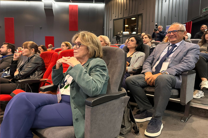 Görselde bir konferans ya da ödül töreninde izleyici koltuklarında oturan insanlar görülüyor. Ön planda yaşlı bir kadın ve yanında takım elbiseli bir erkek dikkat çekiyor. Salonda pek çok kişi etkinliği dikkatle takip ediyor, bazıları gülümserken bazıları düşünceli görünüyor. Arka planda kamera ve teknik ekip de seçilebiliyor.