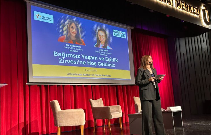 Görselde bir sahne ortamı görülüyor. Arkada kırmızı perde önünde büyük bir ekran bulunuyor. Ekranda “Bağımsız Yaşam ve Eşitlik Zirvesi’ne Hoş Geldiniz” yazısı yer alıyor ve konuşmacıların fotoğrafları ile isimleri gösterilmiş. Sahnenin önünde iki koltuk ve bir sehpa var. Önde ise bir kadın sunucu mikrofon ve tabletle konuşma yapıyor. Mekân Altunizade Kültür ve Sanat Merkezi olarak belirtilmiş.