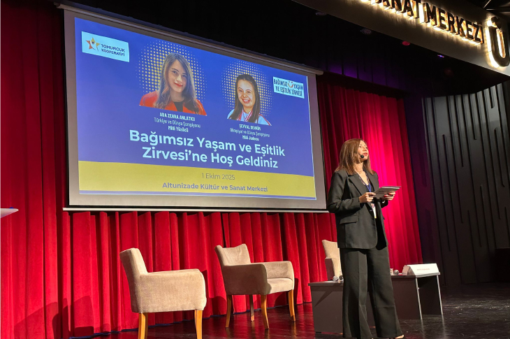 Görselde bir sahne ortamı görülüyor. Arkada kırmızı perde önünde büyük bir ekran bulunuyor. Ekranda “Bağımsız Yaşam ve Eşitlik Zirvesi’ne Hoş Geldiniz” yazısı yer alıyor ve konuşmacıların fotoğrafları ile isimleri gösterilmiş. Sahnenin önünde iki koltuk ve bir sehpa var. Önde ise bir kadın sunucu mikrofon ve tabletle konuşma yapıyor. Mekân Altunizade Kültür ve Sanat Merkezi olarak belirtilmiş.