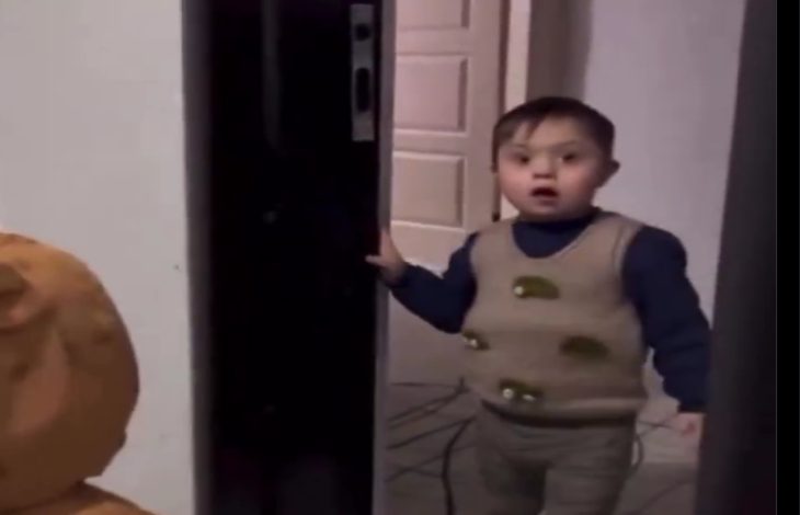 Görselde küçük bir çocuk kapıdan içeri bakıyor. Üzerinde desenli bir süveter ve koyu renk bir kazak var. Yüz ifadesi şaşkınlık ve sevinç karışımı bir duygu yansıtıyor. Arka planda açık bir kapı ve iç mekân detayları görülüyor.