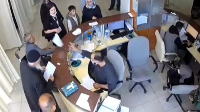 Görselde, yarım daire şeklinde bir danışma masası etrafında çeşitli insanlar toplanmış. Masanın arkasında görevli bir doktor oturuyor ve maske takıyor; elinde bir kağıt tutarken karşısındaki kişilere uzatıyor. Masanın önünde bekleyen kişiler arasında yaşlı bir adam, bir çocuk, kadınlar ve birkaç yetişkin erkek bulunuyor.