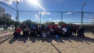 Görselde açık havada, voleybol sahasında bir grup kişi toplu halde poz veriyor. Katılımcıların bir kısmı tekerlekli sandalyede, bazıları ise ayakta duruyor. Arkada tel örgü, ağaçlar ve mavi gökyüzü görünüyor. Güneşli bir günde çekilmiş.