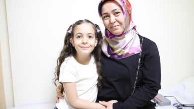 Görselde bir kadın ve küçük kızı yan yana oturmuş, gülümseyerek poz veriyor. Kadın başörtülü, siyah kıyafetli ve kızını kucaklamış. Küçük kız ise beyaz tişört giymiş, saçları iki yandan toplanmış. İkisi de samimi ve mutlu bir şekilde kameraya bakıyor. Ortam sade bir iç mekân, muhtemelen bir hastane odası.