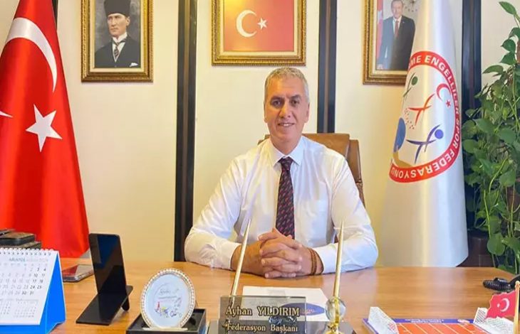 Fotoğrafta bir makam odasında masasında oturan bir erkek görülüyor. Masanın üzerinde isimlik, takvim, plaket ve Türk bayrağı bulunuyor. Arkada Türk bayrağı, bir kurumun bayrağı ve duvarda Atatürk, Türkiye Cumhuriyeti Cumhurbaşkanı ve Türk bayrağı fotoğrafları yer alıyor.