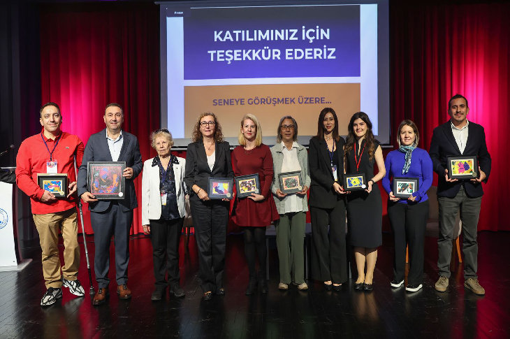 Bir sahnede yan yana duran dokuz kişi, ellerinde plaket veya çerçeveler tutarak poz veriyor. Arkadaki ekranda “Katılımınız için teşekkür ederiz – Seneye görüşmek üzere...” yazısı yer alıyor. Sahne kırmızı perdelerle çevrili.