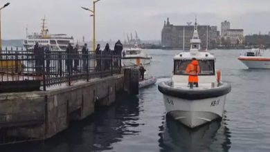 Bir iskelede kurtarma ekipleri ve polisler toplanmış. Denizde bir kurtarma botu ve birkaç tekne bulunuyor. Ekipler suya düşen kişileri kurtarmak için çalışıyor. Arka planda şehir manzarası ve binalar görünüyor.