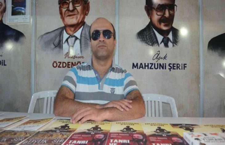 Görselde, masanın arkasında oturan bir erkek bulunuyor. Üzerinde mavi-beyaz çizgili bir tişört ve güneş gözlüğü var. Masada çeşitli kitaplar sergilenmiş. Arka planda duvarda ozan ve yazar portreleri ile isimleri yer alıyor.