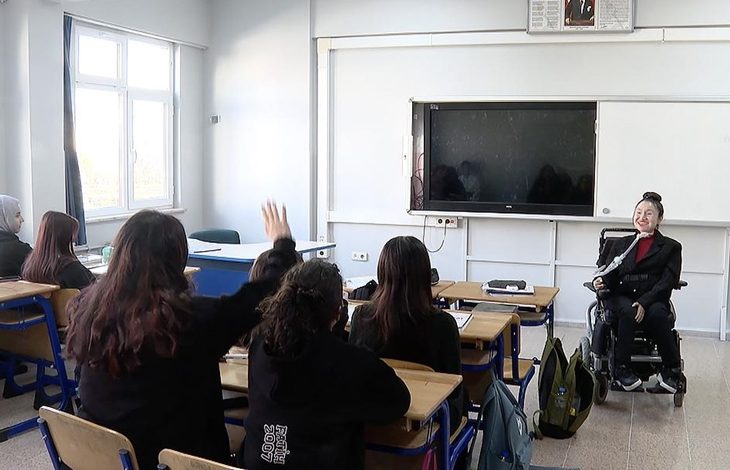 Görselde bir sınıf ortamı görülüyor. Öğretmen tekerlekli sandalyede, öğrencilerine doğru dönmüş şekilde ders anlatıyor. Ön sıradaki bir öğrenci el kaldırmış durumda. Öğretmen gülümserken, öğrenciler dikkatle onu dinliyor. Arka planda büyük bir akıllı tahta ve beyaz tahta yer alıyor.