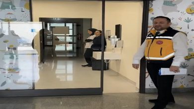 Görselde bir hastane ya da sağlık kurumunun girişinde duran bir görevli görülüyor. Görevli sarı-siyah yelek giymiş ve elinde bir belge tutuyor. Arka planda iki sağlık personeli koridorda ayakta duruyor. Duvarlarda çocuklara yönelik renkli desenler yer alıyor; ortam temiz ve aydınlık bir sağlık tesisi izlenimi veriyor.