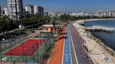 Mersin sahilinde modern bir yürüyüş ve bisiklet yolu yer almakta. Sol tarafta, yeşillikler içinde bir tenis kortu ve çevresinde palmiyelerle süslenmiş park alanı görülüyor. Sağ tarafta ise kayalık bir kıyı boyunca uzanan sahil yürüyüş yolu, denize doğru uzanan bir iskele ile tamamlanıyor. Arka planda ise yüksek apartmanlar ve şehir silueti, Akdeniz'in berrak maviliğiyle birleşerek ferahlatıcı bir atmosfer yaratıyor.
