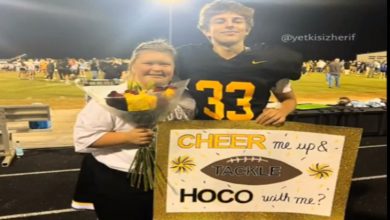 Bir futbol sahasında çekilmiş fotoğrafta, formasıyla bir erkek öğrenci ve çiçek tutan bir kız öğrenci yan yana duruyor. Erkek öğrenci elinde “Cheer me up & tackle HOCO with me?” yazılı süslenmiş bir pano tutuyor. Arkada gece ışıkları altında kalabalık bir grup görünüyor.