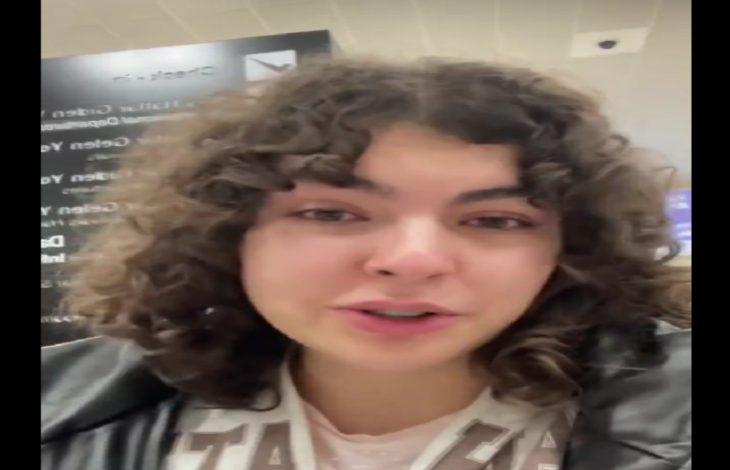 Görselde, kıvırcık saçlı bir kadın kapalı bir alanda kameraya doğru konuşurken görülüyor. Üzerinde açık renkli bir üst ve koyu renkli bir ceket bulunuyor. Arka planda bulanık biçimde yazılar ve tabelalar yer alıyor.