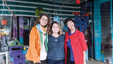 Üç kişi, renkli bir arka plan önünde yan yana durarak gülümsüyor. Üstleri açık bir alanın altında çekilen fotoğrafta, kişiler neşeli bir poz veriyor. Arka planda bitkiler ve dekoratif objeler görülüyor.