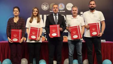 Görselde beş kişi yan yana duruyor. Ellerinde kırmızı renkli dosyalar tutuyorlar. Ortadaki kişi takım elbiseli, diğer dört kişi ise daha sade kıyafetli. Arka planda "DUY BENİ" yazısı ve bazı kurum logoları yer almakta. Ön tarafta ise süsleme amaçlı balonlar bulunuyor. Bu görüntü, resmi bir tören ya da proje tanıtımı kapsamında çekilmiş gibi görünüyor.