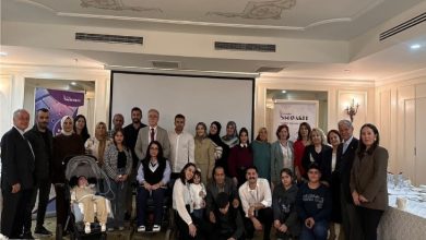 Görselde, bir toplantı salonunda kalabalık bir grup hatıra fotoğrafı çektiriyor. Katılımcılar farklı yaş ve gruplardan oluşuyor; aralarında tekerlekli sandalye kullanan ve bebek arabasıyla gelen kişiler de var. Arka planda bir projeksiyon perdesi ve etkinlik afişleri görülüyor. Ortam resmi ve samimi bir buluşma havasında.