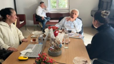 Görselde bir masa etrafında oturmuş üç kişi sohbet ediyor. Masada çay bardakları, laptop, su şişesi ve çiçek saksıları bulunuyor. Arka planda pencereden gün ışığı geliyor ve bir kişi daha oturmuş şekilde görülüyor. Ortam samimi bir ofis ya da toplantı odası havasında.