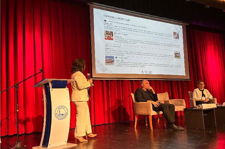 Görselde bir konferans salonunda sunum yapıldığı görülüyor. Kırmızı perde önünde büyük bir projeksiyon ekranında “İBB Engelli Hizmetleri” başlıklı bir sunum yansıtılmış. Sol tarafta kürsüde mikrofonla bir kadın sunum yaparken, sahnenin ortasında iki koltukta oturan konuşmacılar yer alıyor. Katılımcılar dikkatle sunumu takip ediyor.