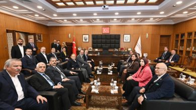 Geniş, resmi bir toplantı salonunda büyük bir masa etrafında oturan kalabalık bir heyet görülüyor. Takım elbiseli erkekler ve birkaç kadın, protokol düzeninde karşılıklı sıralanmış. Arka planda Türk bayrağı, makam koltuğu ve duvara asılı portreler yer alıyor. Atmosfer ciddi ve resmi.