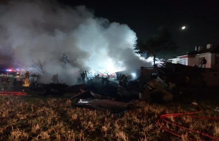 Görselde gece saatlerinde çıkan bir yangının ardından yoğun dumanın gökyüzüne yükseldiği bir alan görülüyor. İtfaiye ekipleri ve görevliler, alevlerin söndürüldüğü enkazın arasında çalışıyor. Önde yanmış malzemeler ve yıkıntılar yer alırken, arka planda itfaiye araçlarının ışıkları dumanın içinde parlıyor. Sahne, yangının büyük hasara yol açtığı ve müdahalenin hâlâ sürdüğü dramatik bir durumu yansıtıyor.