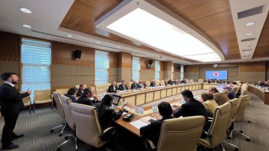 Görselde, büyük bir toplantı salonunda uzun bir masa etrafında toplanmış çok sayıda kişi görülüyor. Katılımcılar resmi kıyafetler içinde, toplantı sırasında notlar alıyor ve konuşmaları dinliyor. Salon modern bir tasarıma sahip; duvarda Türkiye Büyük Millet Meclisi logosu bulunan bir ekran yer alıyor.