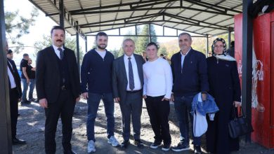 Görselde, bir grup kişi açık alanda, üzeri kapalı bir yapının altında poz veriyor. Ortada takım elbiseli bir erkek bulunuyor; yanında casual giyimli gençler ve bir kadın yer alıyor. Arka planda ağaçlar ve birkaç kişi daha görünüyor. Ortam samimi ve ziyaret havasında.