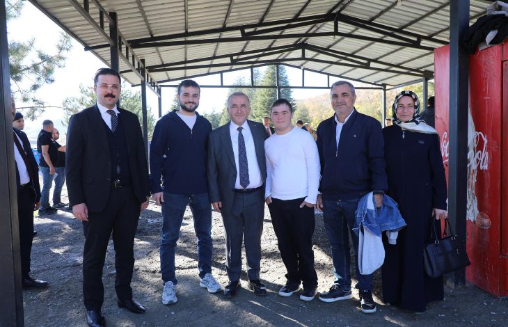 Görselde, bir grup kişi açık alanda, üzeri kapalı bir yapının altında poz veriyor. Ortada takım elbiseli bir erkek bulunuyor; yanında casual giyimli gençler ve bir kadın yer alıyor. Arka planda ağaçlar ve birkaç kişi daha görünüyor. Ortam samimi ve ziyaret havasında.